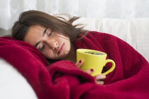 Sick-looking woman wrapped up in red blanket and holding yellow cup